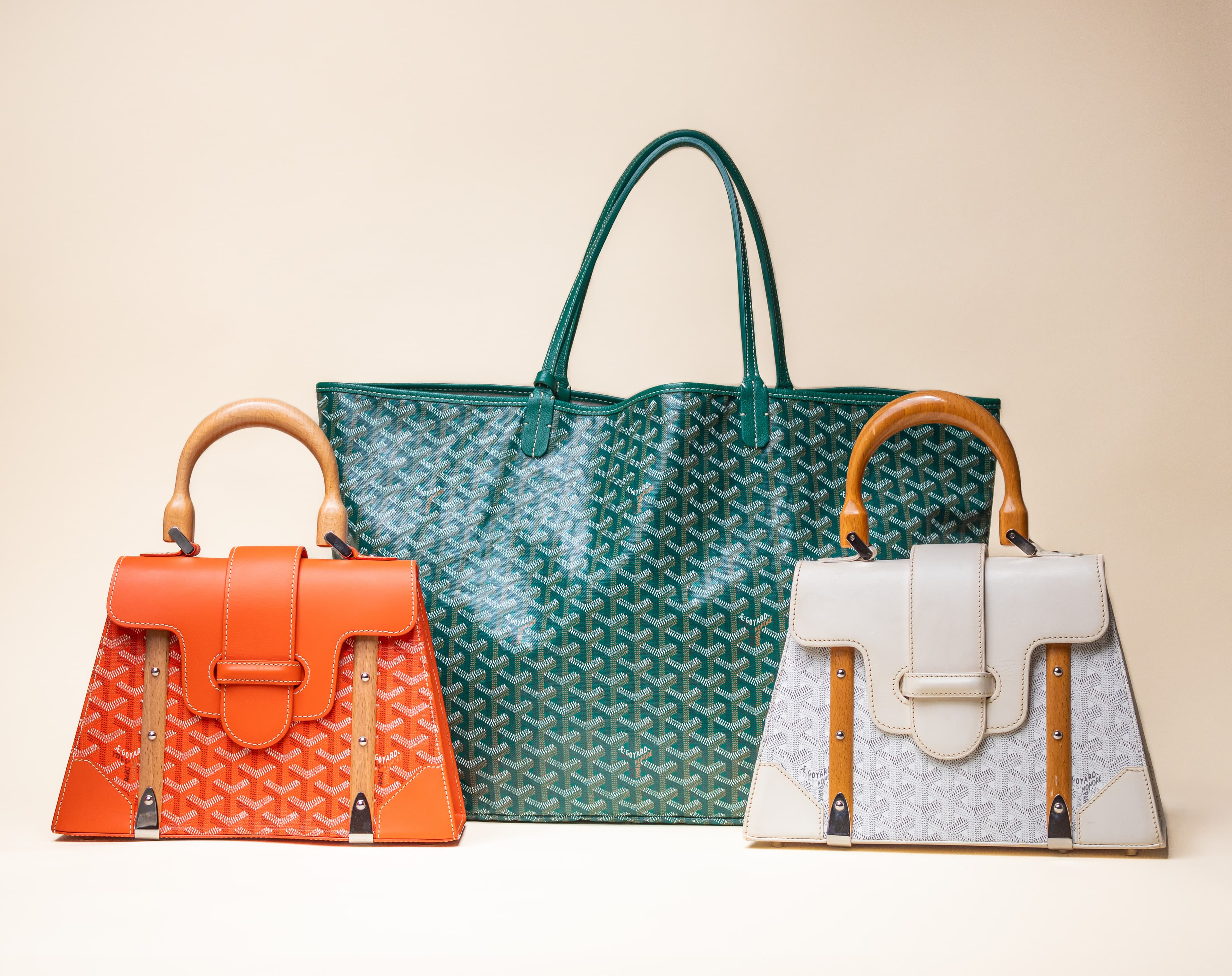 Three designer handbags: a large green tote with pattern, flanked by orange and white bags with wooden handles.